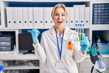 Young caucasian woman working at scientist laboratory holding test tubes pointing thumb up to the side smiling happy with open mouth