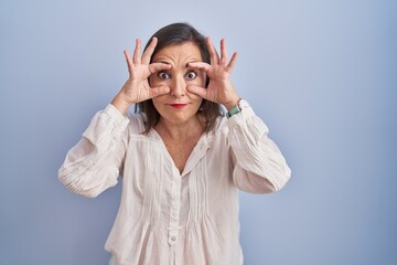 Middle age hispanic woman standing over blue background trying to open eyes with fingers, sleepy and tired for morning fatigue