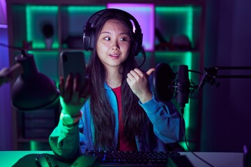 Young asian woman playing video games with smartphone smiling with happy face looking and pointing to the side with thumb up.