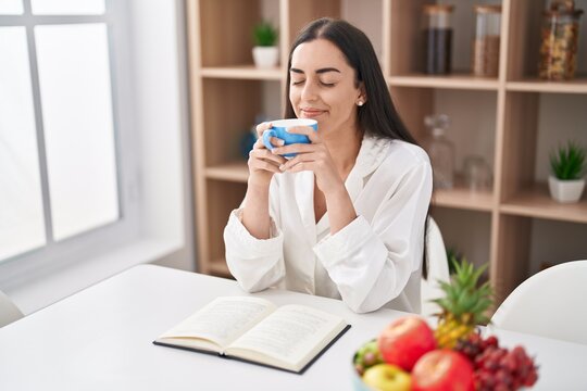 Young hispanic woman reading book drinking coffee at home