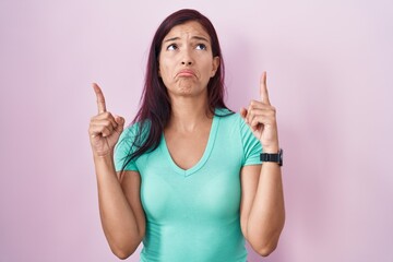 Young hispanic woman standing over pink background pointing up looking sad and upset, indicating...