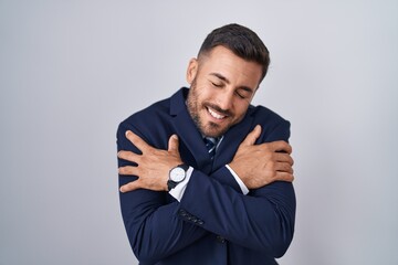 Handsome hispanic man wearing suit and tie hugging oneself happy and positive, smiling confident. self love and self care