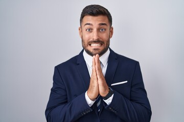 Handsome hispanic man wearing suit and tie praying with hands together asking for forgiveness smiling confident.