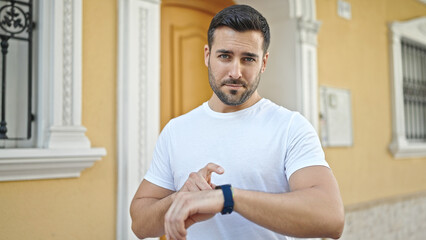 Young hispanic man looking watch with relaxed expression at street