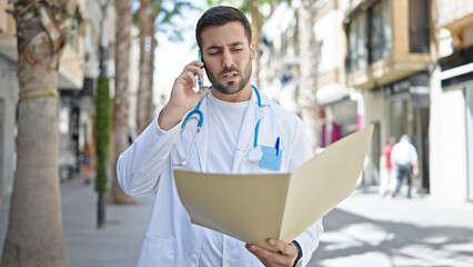 Young hispanic man doctor reading medical report talking on smartphone at street