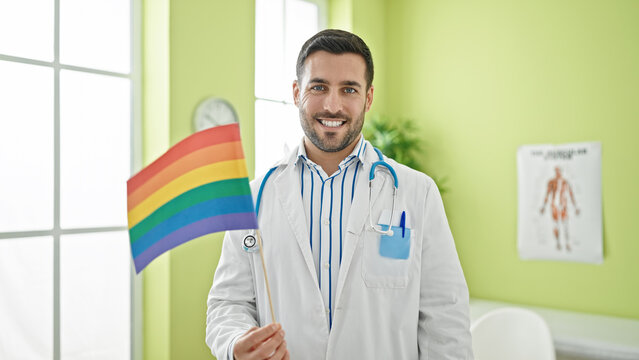 Young hispanic man doctor smiling confident holding lgbtq flag at clinic