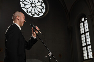 Mature pastor in black suit speaking prayer in microphone while standing in old baptist church