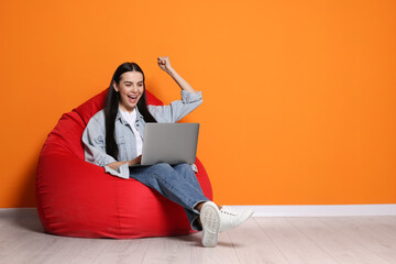 Cheerful woman with laptop sitting on beanbag chair near orange wall, space for text