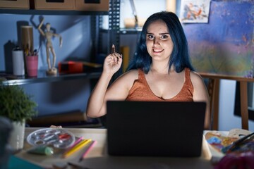 Fototapeta na wymiar Young modern girl with blue hair sitting at art studio with laptop at night with a big smile on face, pointing with hand and finger to the side looking at the camera.