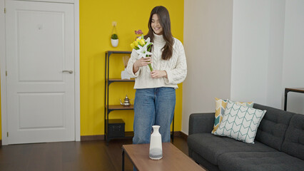 Young beautiful hispanic woman putting flowers on pot at home