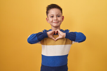 Little hispanic boy standing over yellow background smiling in love doing heart symbol shape with...