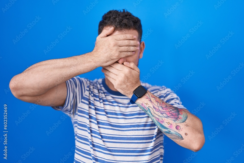 Sticker young hispanic man standing over blue background covering eyes and mouth with hands, surprised and s