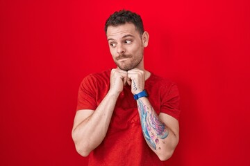 Young hispanic man standing over red background laughing nervous and excited with hands on chin...
