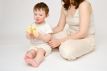 Happy baby with mother eating banana on studio white background. Portrait of a smiling child with mom snacking on fruits. Kid about two years old (one year nine months)