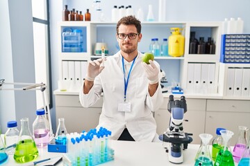 Young hispanic man working at scientist laboratory holding apple relaxed with serious expression on face. simple and natural looking at the camera.