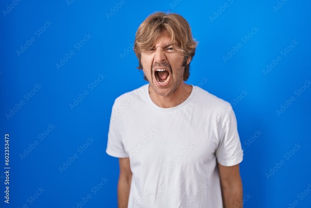 Poster middle age man standing over blue background angry and mad screaming frustrated and furious, shoutin