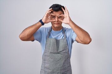 Hispanic young man wearing apron over white background trying to open eyes with fingers, sleepy and tired for morning fatigue