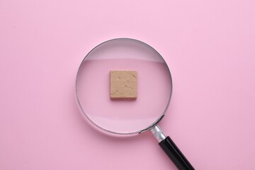 Looking at wooden cube through magnifying glass on pink background, top view