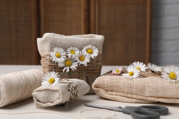 Gifts packed in burlap fabric, beautiful chamomiles, ropes, spool of threads and scissors on white...