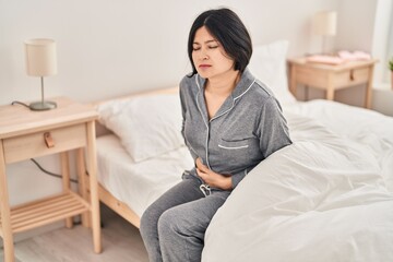 Obraz premium Young chinese woman suffering for menstrual pain sitting on bed at bedroom