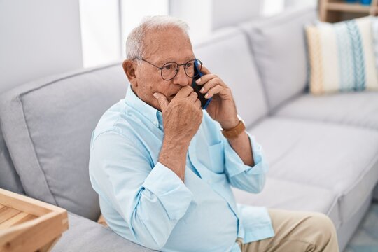 Senior grey-haired man talking on smartphone sitting on sofa at home