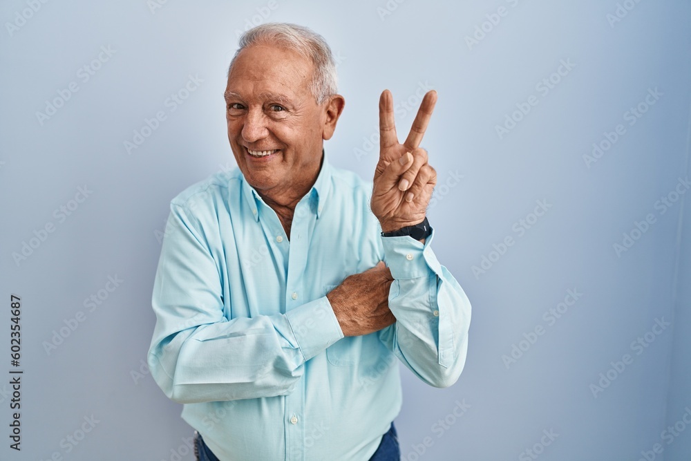 Sticker Senior man with grey hair standing over blue background smiling with happy face winking at the camera doing victory sign. number two.