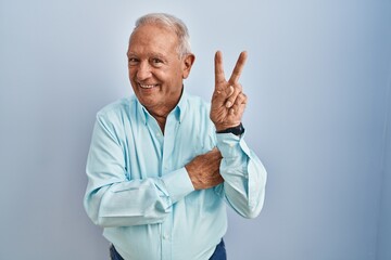 Senior man with grey hair standing over blue background smiling with happy face winking at the camera doing victory sign. number two.