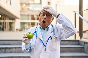 Senior doctor with grey hair holding healthy green apple stressed and frustrated with hand on head,...