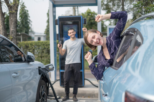 Rusain woman smile to camera and fist her hand with happiness in her vacation, hand happy by save money with electral car fluel station with blur happy her boy friend.