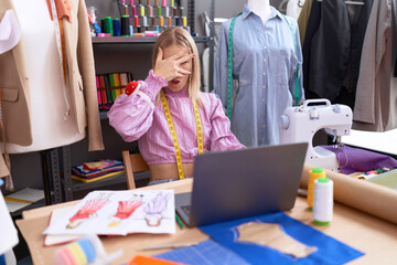Young caucasian woman dressmaker designer using laptop peeking in shock covering face and eyes with...