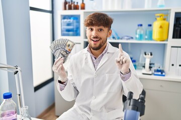 Arab man with beard working at scientist laboratory holding money surprised with an idea or question pointing finger with happy face, number one