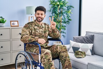 Fototapeta premium Arab man wearing camouflage army uniform sitting on wheelchair showing and pointing up with fingers number two while smiling confident and happy.