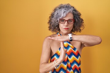 Middle age woman with grey hair standing over yellow background doing time out gesture with hands,...