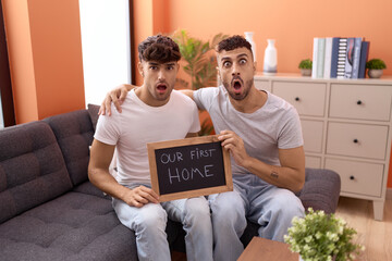 Homosexual gay couple holding blackboard with first home text afraid and shocked with surprise and...