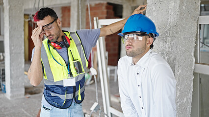 Two men builder and architect standing together with stressed expression at construction site