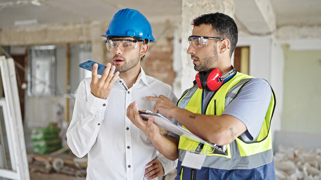 Two men builders and architect writing on document sending voice message by smartphone at construction site