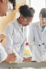 Vertical portrait of teen black schoolgirl wearing lab coat in science class and doing experiments