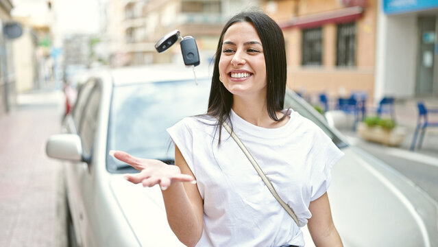 Young beautiful hispanic woman holding key standing by car at street