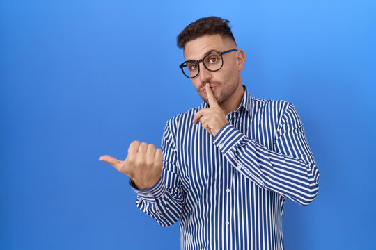 Hispanic man with beard wearing glasses asking to be quiet with finger on lips pointing with hand to the side. silence and secret concept.
