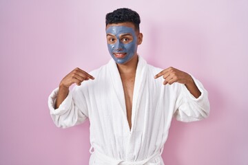 Young hispanic man wearing beauty face mask and bath robe looking confident with smile on face,...