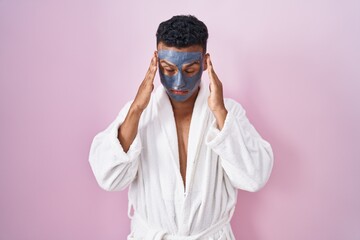 Young hispanic man wearing beauty face mask and bath robe with hand on head, headache because stress. suffering migraine.