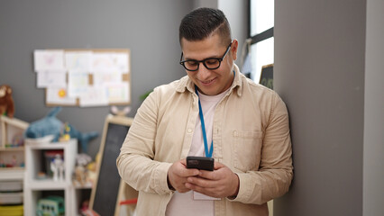 Young hispanic man preschool teacher using smartphone at kindergarten