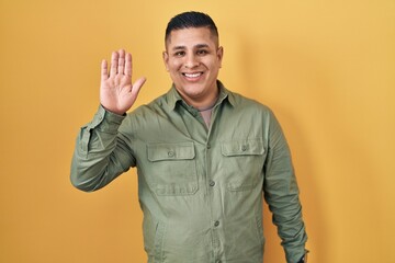 Hispanic young man standing over yellow background waiving saying hello happy and smiling, friendly welcome gesture