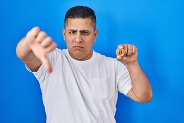 Hispanic young man holding virtual currency bitcoin with angry face, negative sign showing dislike with thumbs down, rejection concept