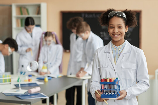 Waist up portrait of black teen schoolgirl wearing lab coat in science class and smiling at camera with group of children in background, copy space