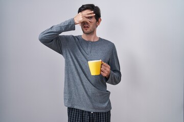 Young hispanic man wearing pajama drinking a cup of coffee peeking in shock covering face and eyes...