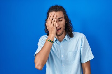 Young hispanic man standing over blue background yawning tired covering half face, eye and mouth with hand. face hurts in pain.
