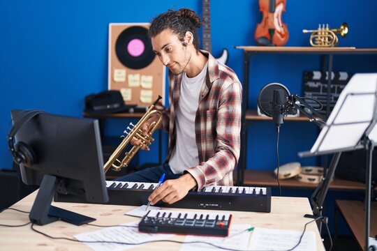 Young hispanic man musician composing song holding trumpet at music studio