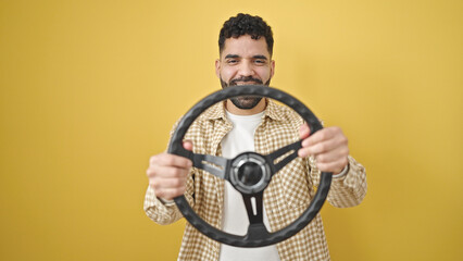 Young hispanic man smiling confident using steering wheel as a driver over isolated yellow background