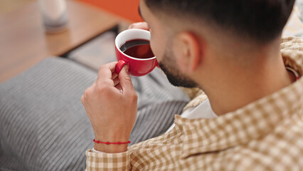 Young hispanic man drinking coffee sitting on sofa at h